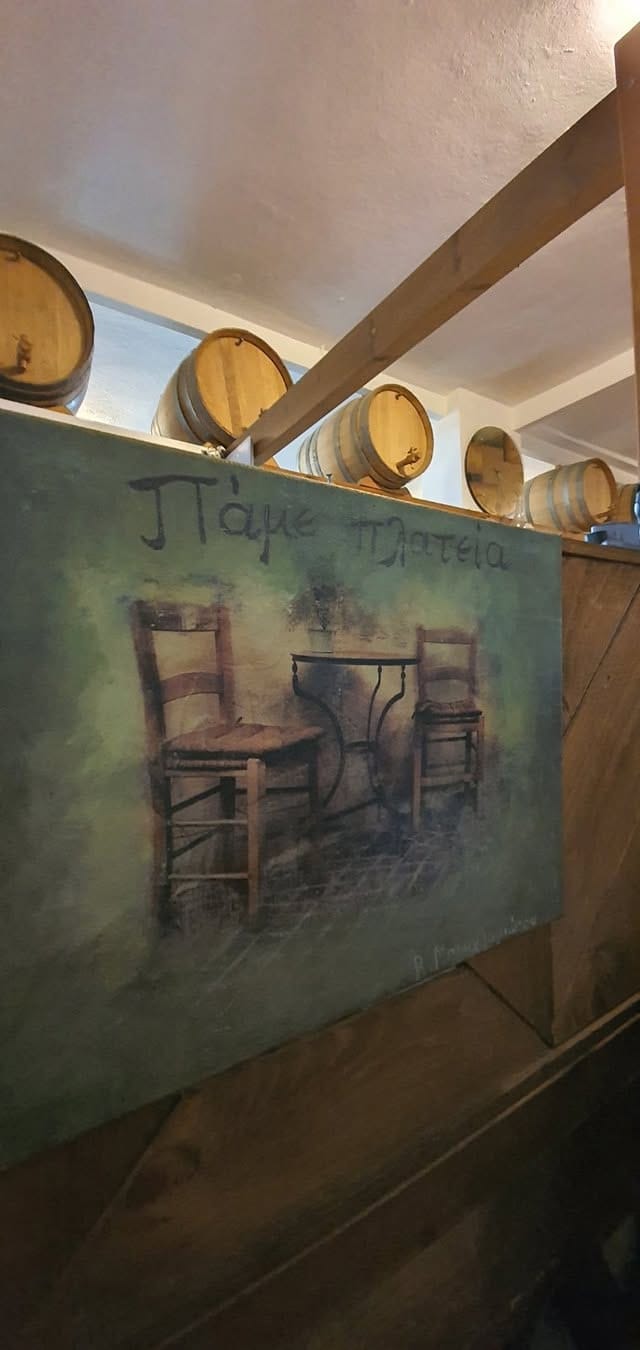 Ξύλινα βαρέλια κρασιού σε ράφι πάνω από χειροποίητο ζωγραφικό πίνακα με το όνομα «Πάμε πλατεία», στον εσωτερικό χώρο του μαγαζιού.