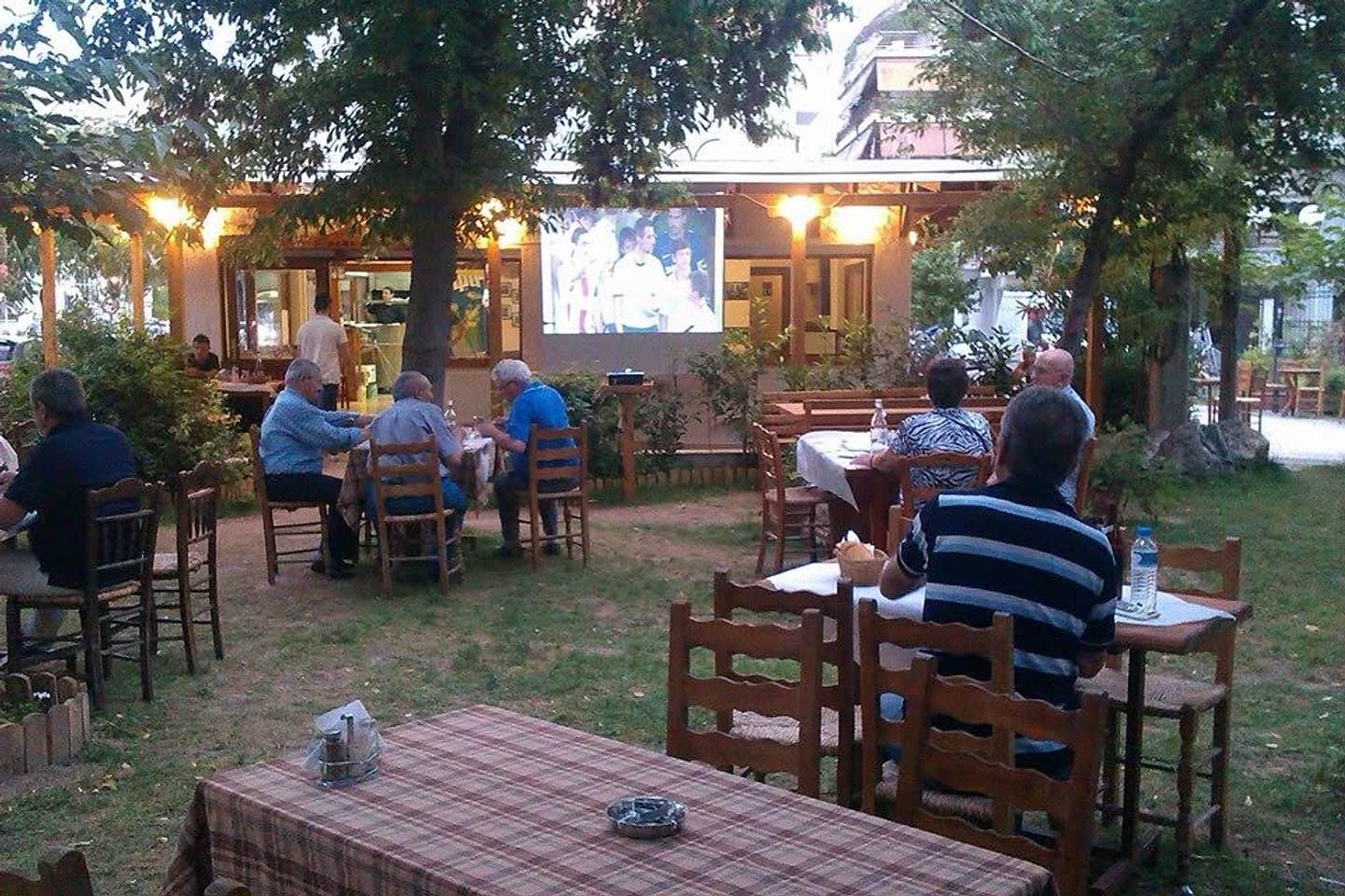 Ο εξωτερικός χώρος του Πάμε Πλατεία στο πάρκο: ξύλινες καρέκλες, καρό τραπεζομάντηλα και παρέες κάτω από τα δέντρα, ένα καλοκαιρινό βράδυ.