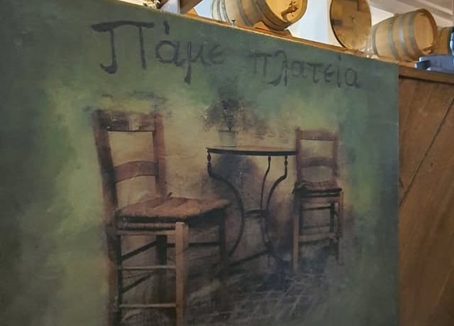Χειροποίητος ζωγραφικός πίνακας με ψάθινες καρέκλες και σιδερένιο τραπεζάκι, με το όνομα «Πάμε πλατεία» γραμμένο στο χέρι.